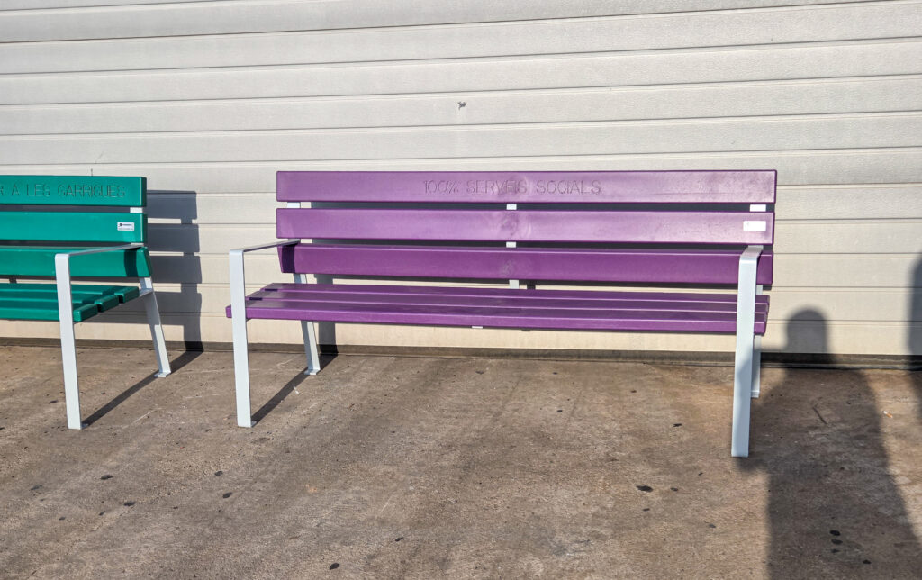 Dos bancos de colores (morado y verde) con patas metálicas instalados frente a pared panelada.